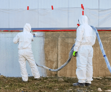 Двама мъже в защитни костюми извършват ремонтни услуги чрез пръскане на бетонна стена.