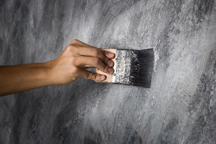 Ръка, която държи четка, нанася черна боя върху текстурирана сива стена.
